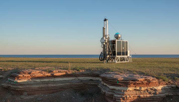 Мичиган: старт разведки природных запасов водорода Современная буровая установка для геологической разведки в поле Мичигана, на заднем плане видны скальные породы и большое озеро.